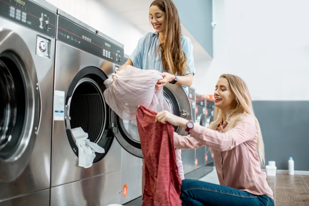 Two cheerful girlfriends having fun loading clothes into the washing machine in the self-service laundry