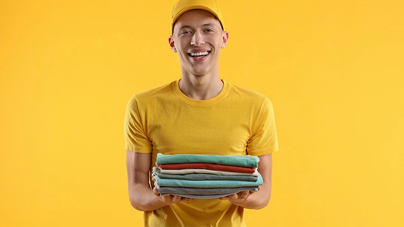 Dry-cleaning delivery. Happy courier holding folded clothes on a yellow background