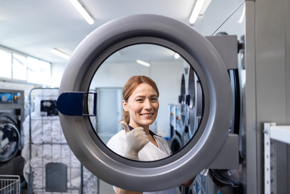 Female laundry worker standing by industrial washing machine and holding thumbs up in dry cleaning service.