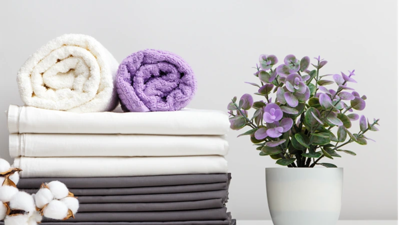 Stack of bed linen, sheets and towels with cotton branch and potted plant on table