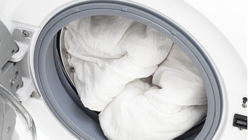 Washing bedding and duvet in washing machine. Laundry care for white blankets and comforter. Household cleaning concept. Clean bed linen inside modern washer close up detail